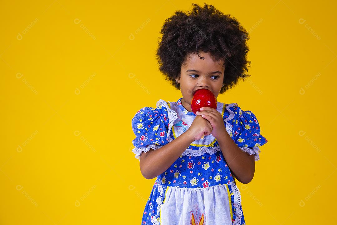 Adorável menina negra vestida com roupas de festa de junho comendo a maçã do amor. Sobremesa de maçã