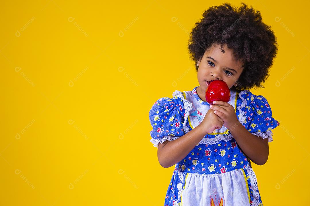 Adorável menina negra vestida com roupas de festa de junho comendo a maçã do amor. Sobremesa de maçã