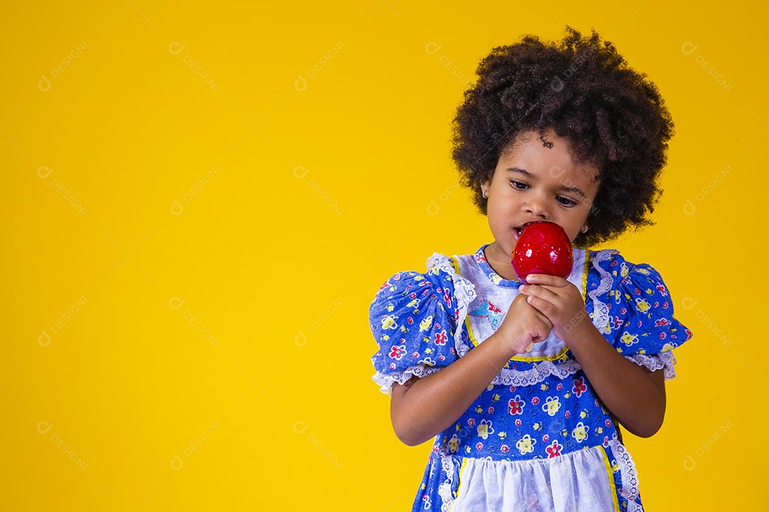 Adorável menina negra vestida com roupas de festa de junho comendo a maçã do amor. Sobremesa de maçã