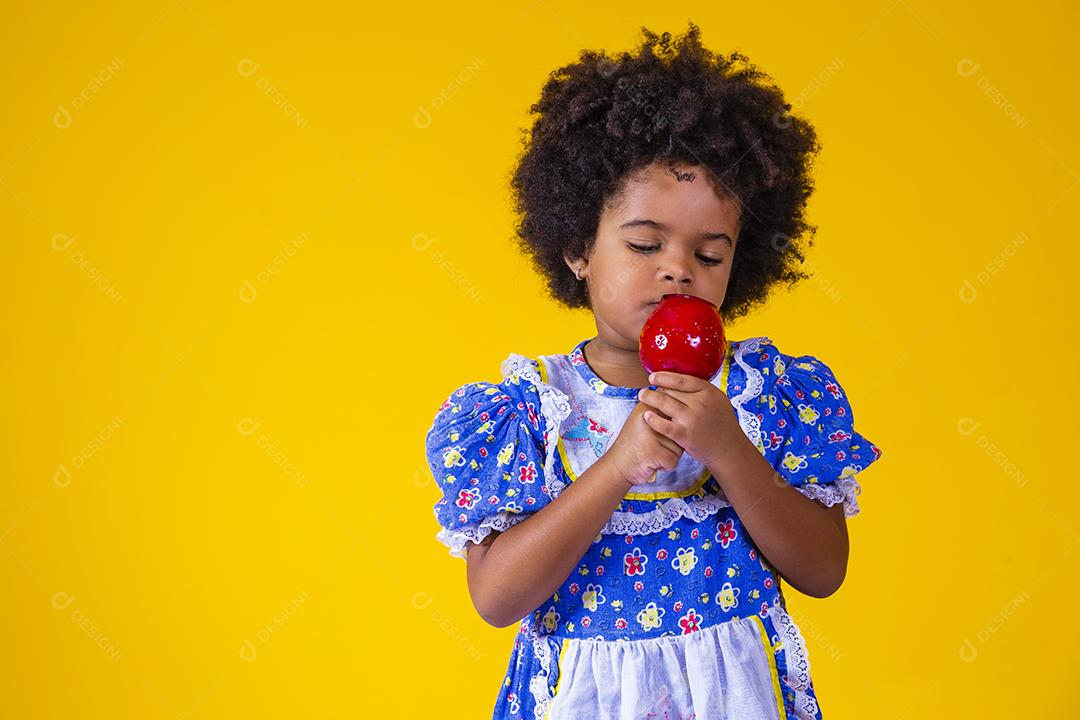 Adorável menina negra vestida com roupas de festa de junho comendo a maçã do amor. Sobremesa de maçã