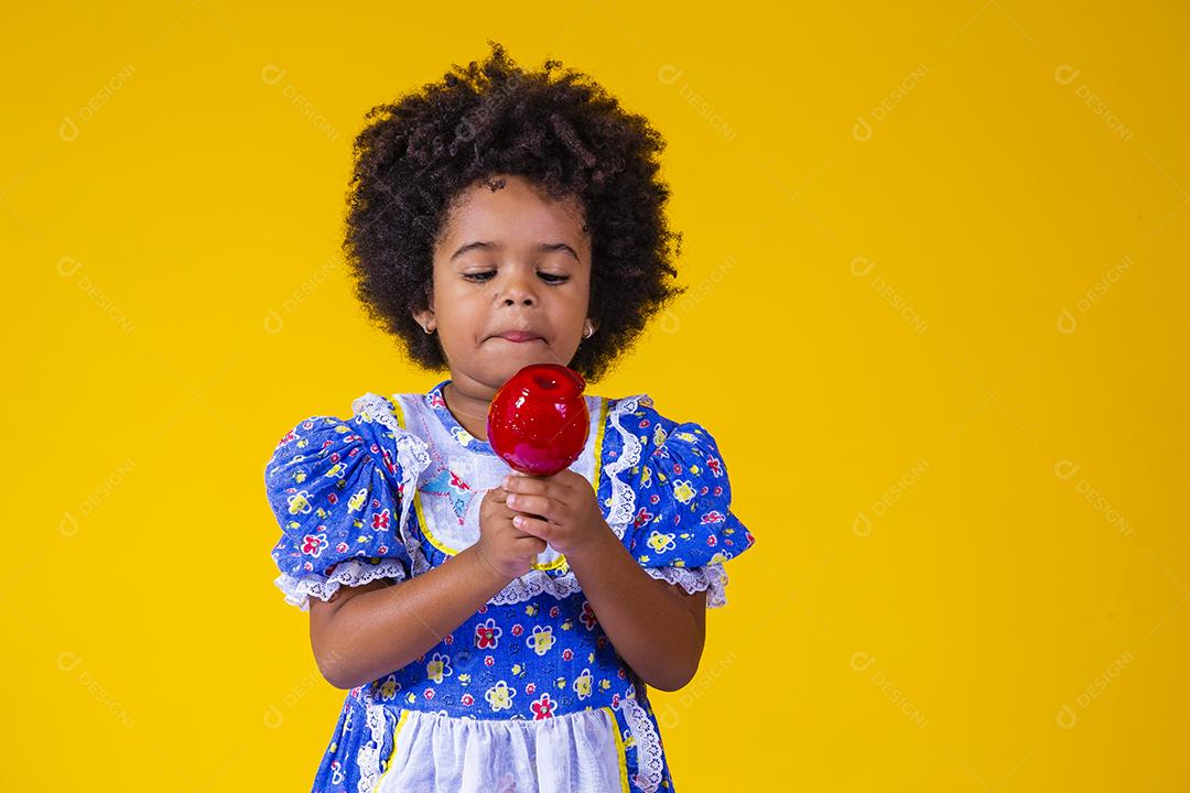 Adorável menina negra vestida com roupas de festa de junho comendo a maçã do amor. Sobremesa de maçã