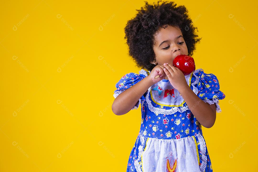 Adorável menina negra vestida com roupas de festa de junho comendo a maçã do amor. Sobremesa de maçã