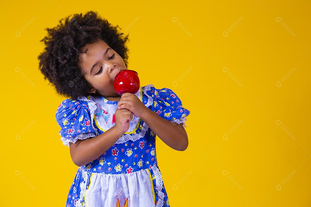 Adorável menina negra vestida com roupas de festa de junho comendo a maçã do amor. Sobremesa de maçã