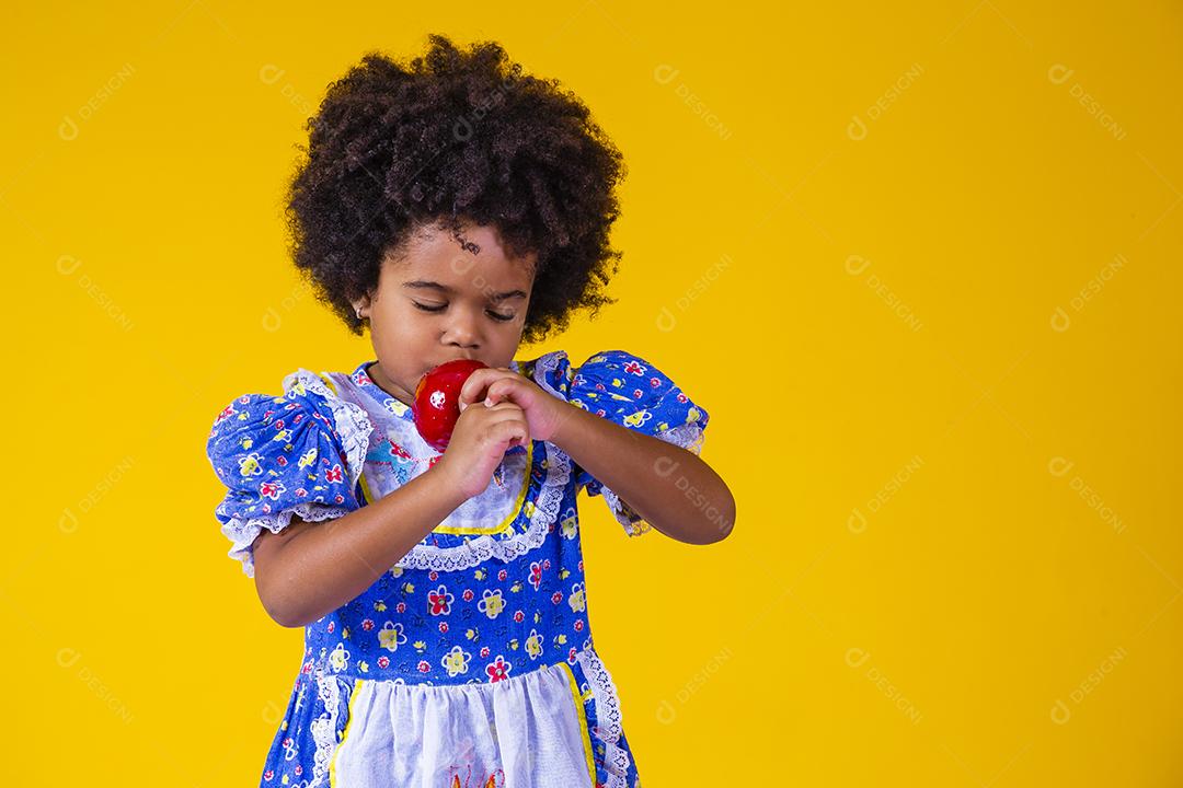 Adorável menina negra vestida com roupas de festa de junho comendo a maçã do amor. Sobremesa de maçã