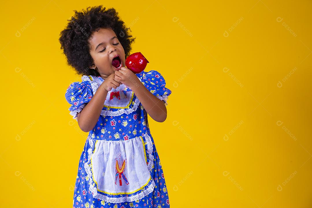 Adorável menina negra vestida com roupas de festa de junho comendo a maçã do amor. Sobremesa de maçã