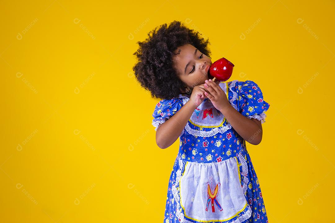 Adorável menina negra vestida com roupas de festa de junho comendo a maçã do amor. Sobremesa de maçã