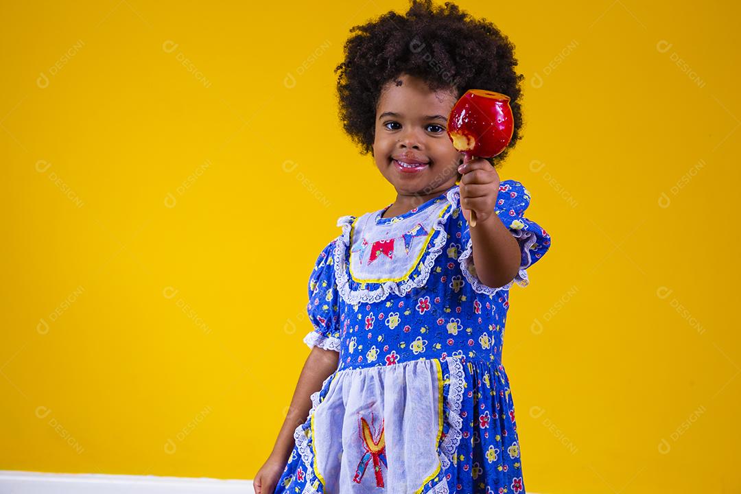 Adorável menina negra vestida com roupas de festa de junho comendo a maçã do amor. Sobremesa de maçã