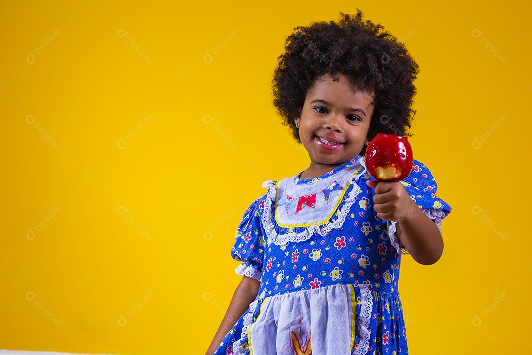 Adorável menina negra vestida com roupas de festa de junho comendo a maçã do amor. Sobremesa de maçã