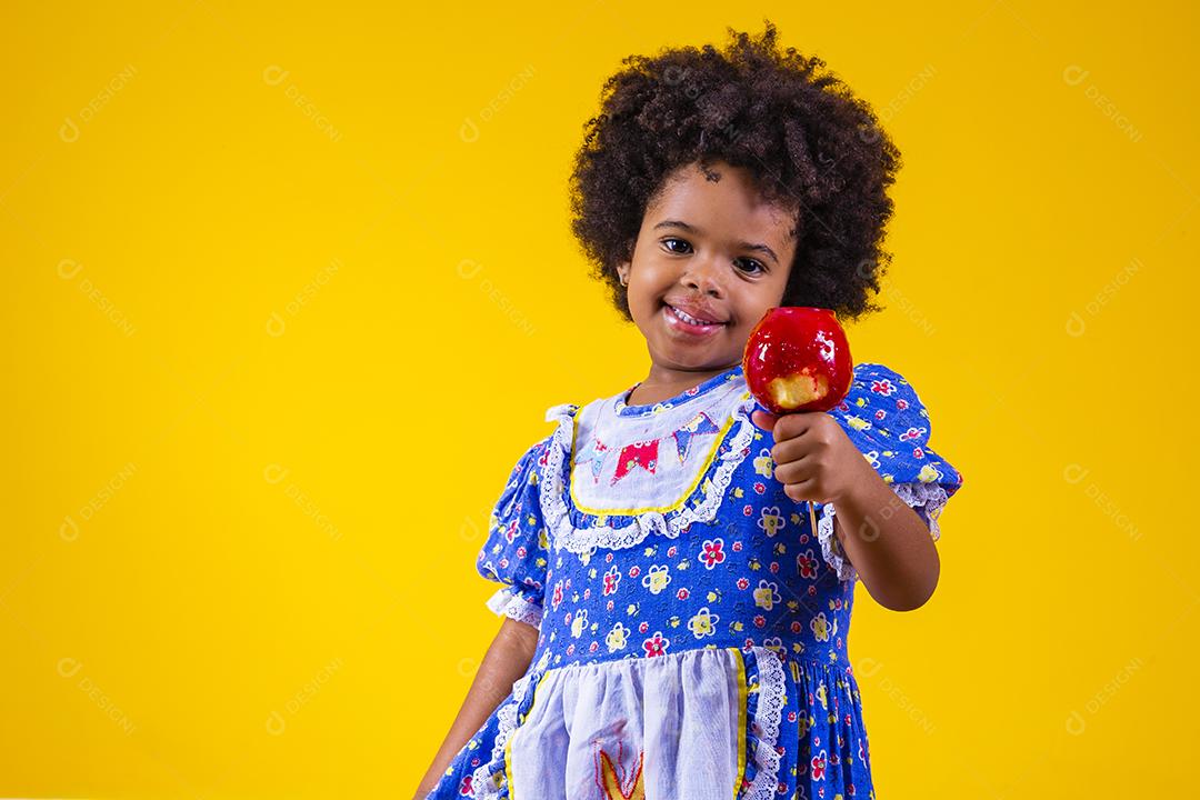 Adorável menina negra vestida com roupas de festa de junho comendo a maçã do amor. Sobremesa de maçã