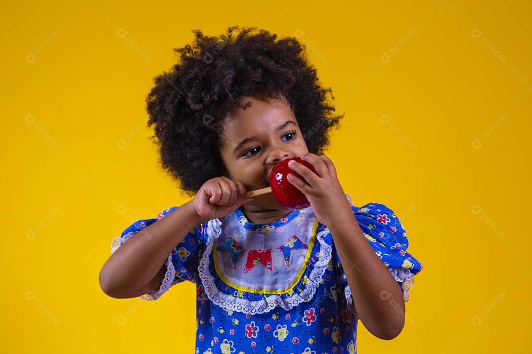 Adorável menina negra vestida com roupas de festa de junho comendo a maçã do amor. Sobremesa de maçã