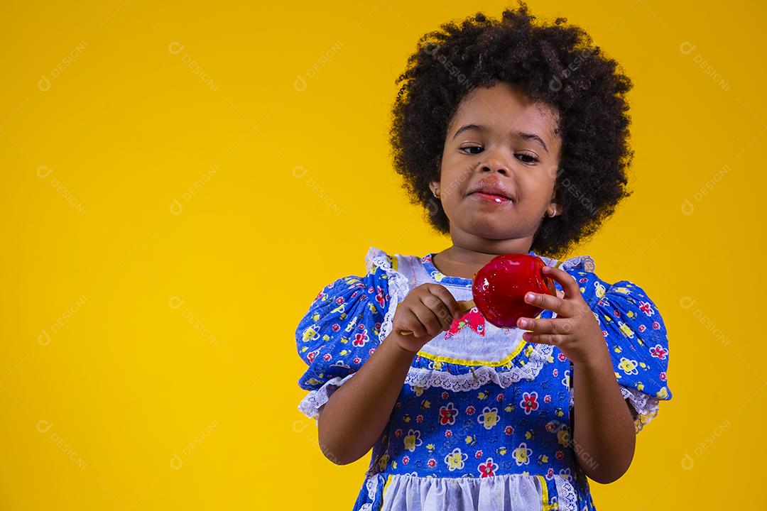 Adorável menina negra vestida com roupas de festa de junho comendo a maçã do amor. Sobremesa de maçã