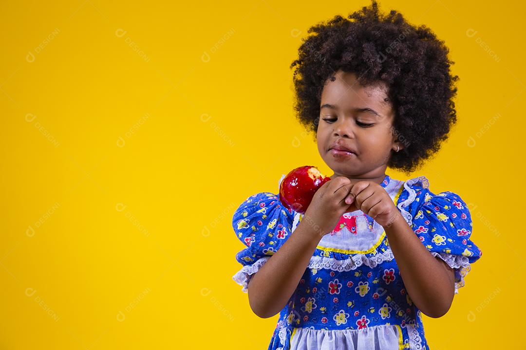 Adorável menina negra vestida com roupas de festa de junho comendo a maçã do amor. Sobremesa de maçã