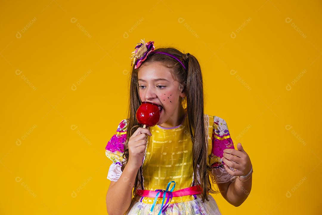 Linda garota com roupas de festa de junho comendo a maçã do amor. Sobremesa de maçã coberta de caramelo vermelho