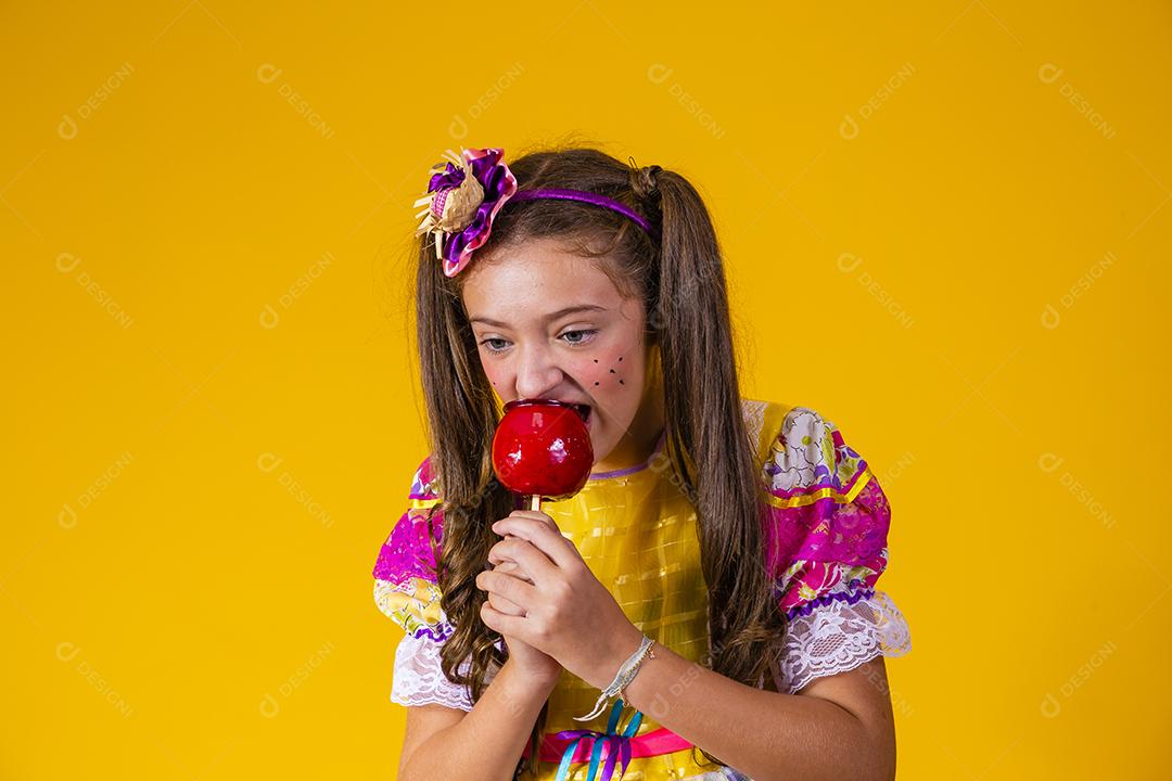 Linda garota com roupas de festa de junho comendo a maçã do amor. Sobremesa de maçã coberta de caramelo vermelho