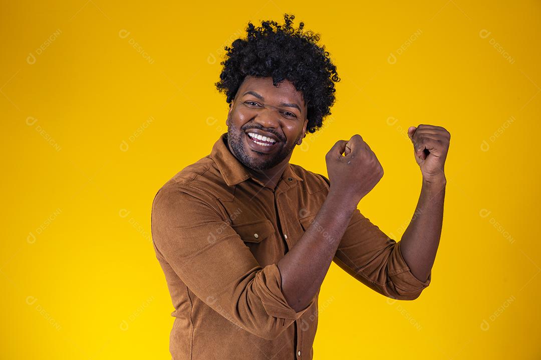 Homem afro comemorando grande sorte e sucesso olhando para câmera posando no estúdio sobre fundo amarelo