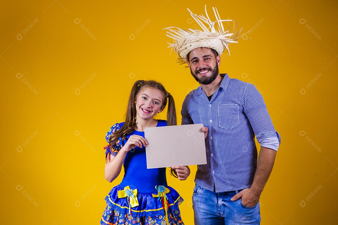 Pai e filha vestidos com roupas de festa junina segurando uma placa