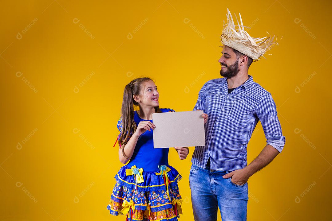 Pai e filha vestidos com roupas de festa junina segurando uma placa