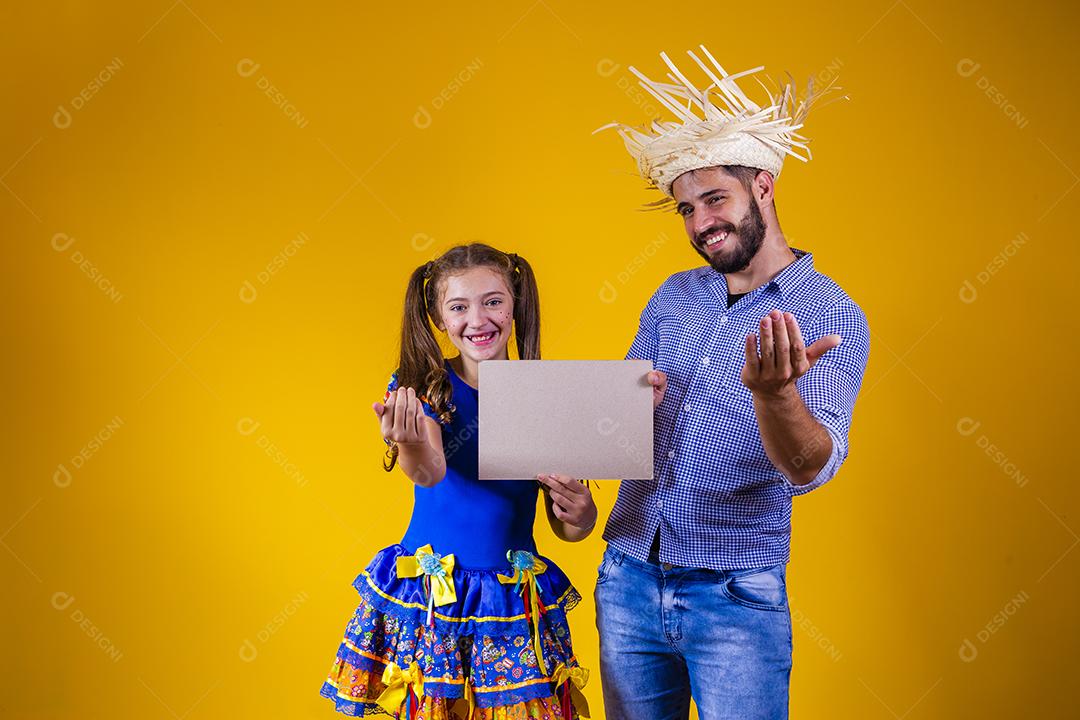 Pai e filha vestidos com roupas de festa junina segurando uma placa
