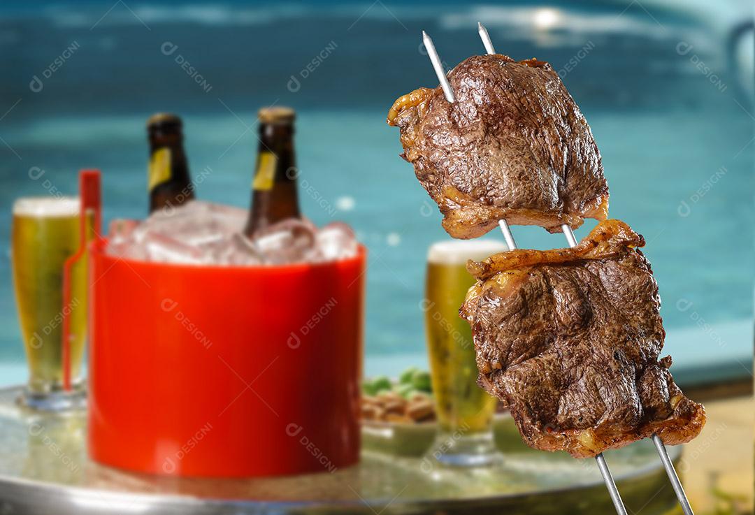 Picanha de churrasco brasileiro em espetos com cervejas turva e piscina ao fundo.