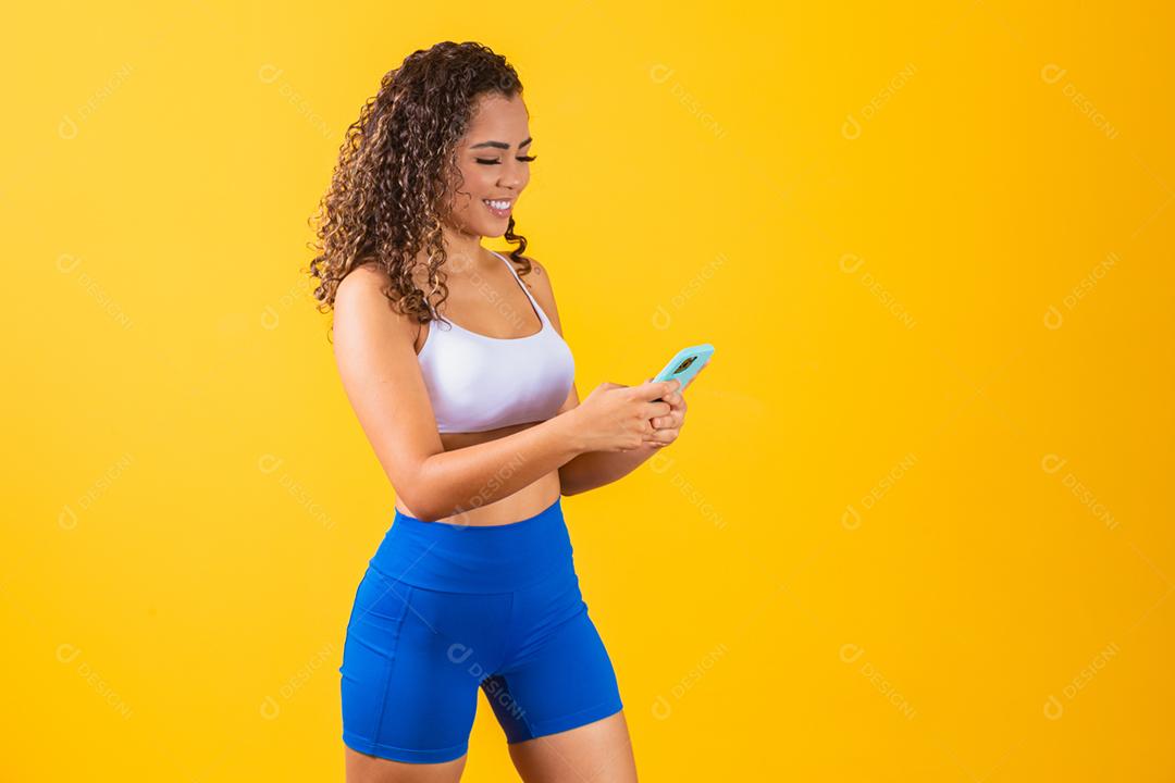 Mulher influenciadora em roupa de fitness usando telefone celular em amarelo ba