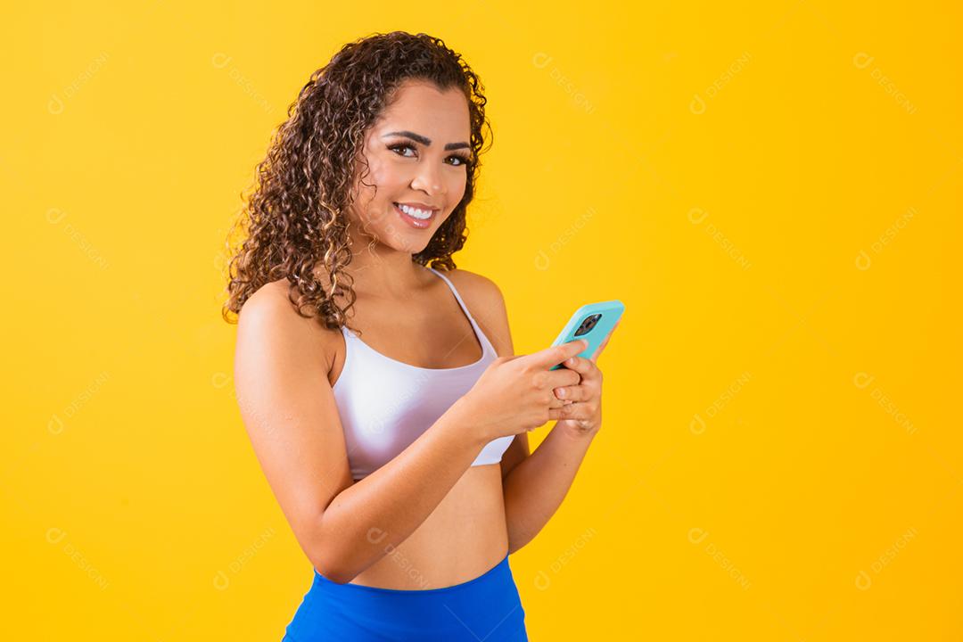 Mulher influenciadora em roupa de fitness usando telefone celular em amarelo ba