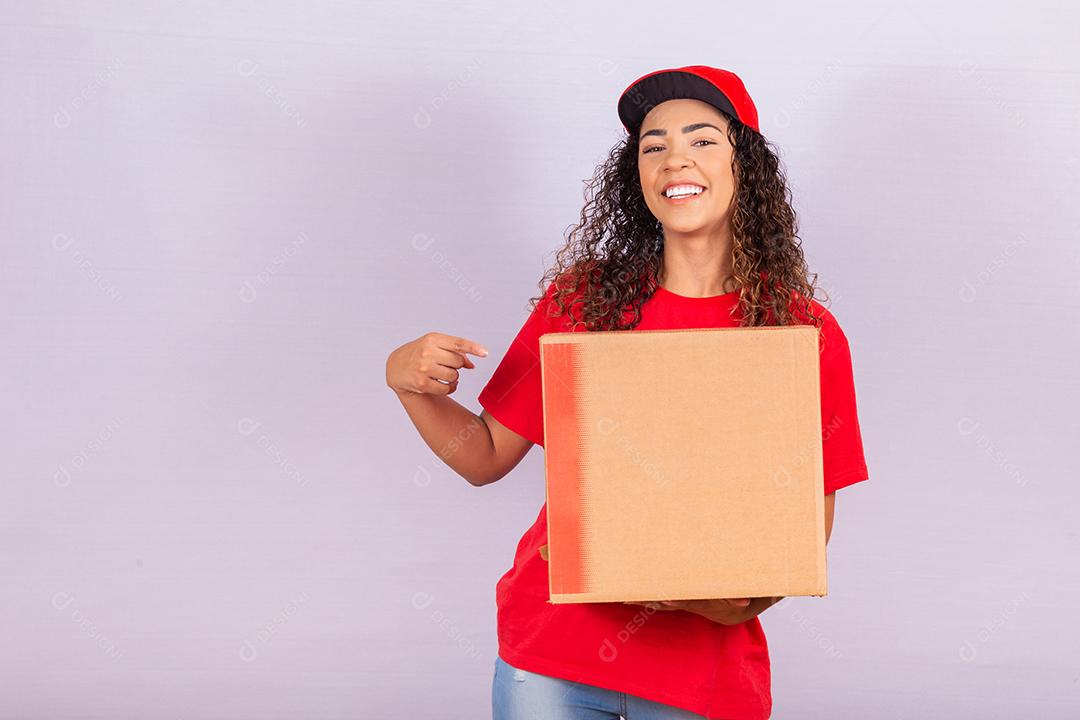 Mulher de entrega afro em um boné segurando uma caixa sobre fundo branco, apontando para uma caixa.
