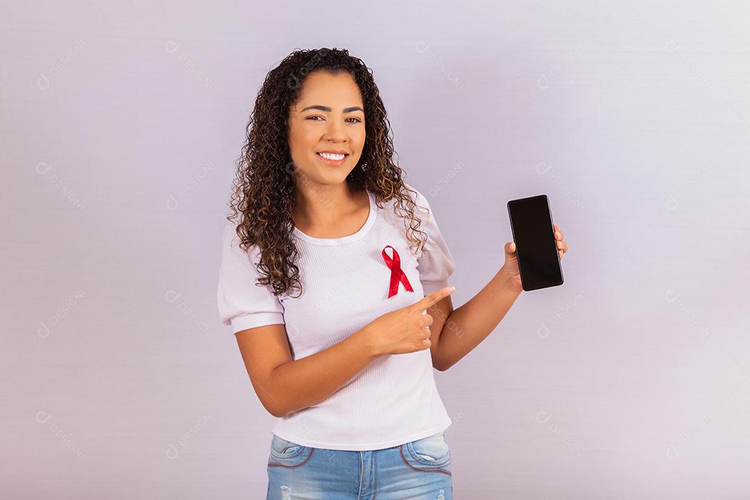 O conceito do Dia Mundial da AIDS. Jovem segura smartphone com tela em branco e fita vermelha na camiseta