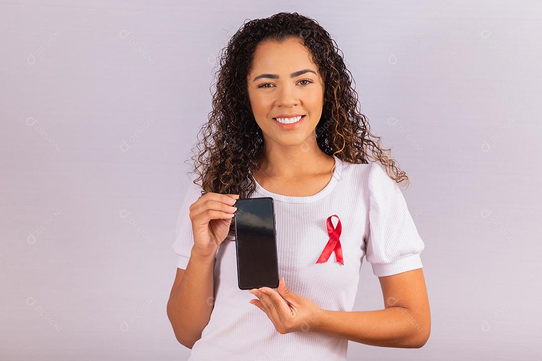 O conceito do Dia Mundial da AIDS. Jovem segura smartphone com tela em branco e fita vermelha na camiseta