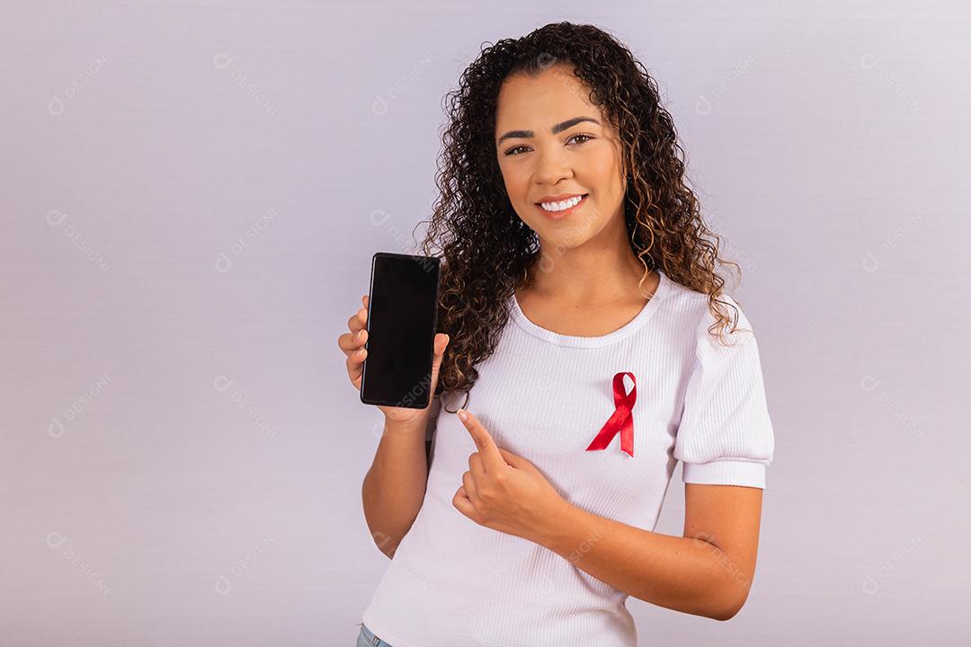 O conceito do Dia Mundial da AIDS. Jovem segura smartphone com tela em branco e fita vermelha na camiseta