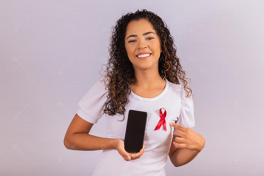 O conceito do Dia Mundial da AIDS. Jovem segura smartphone com tela em branco e fita vermelha na camiseta