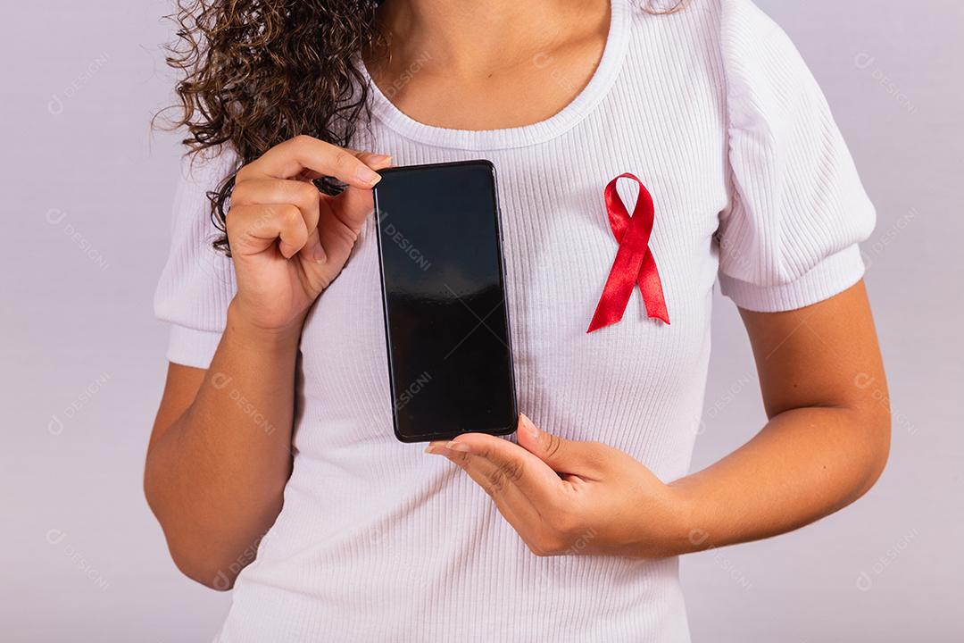 O conceito do Dia Mundial da AIDS. Jovem segura smartphone com tela em branco e fita vermelha na camiseta