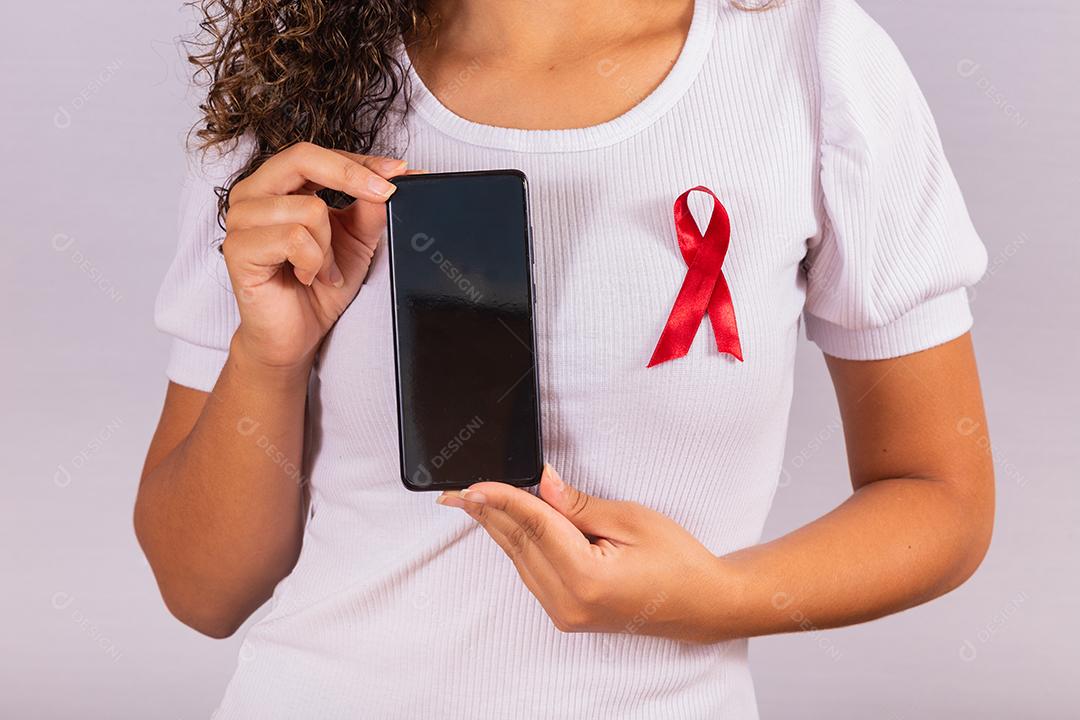 O conceito do Dia Mundial da AIDS. Jovem segura smartphone com tela em branco e fita vermelha na camiseta