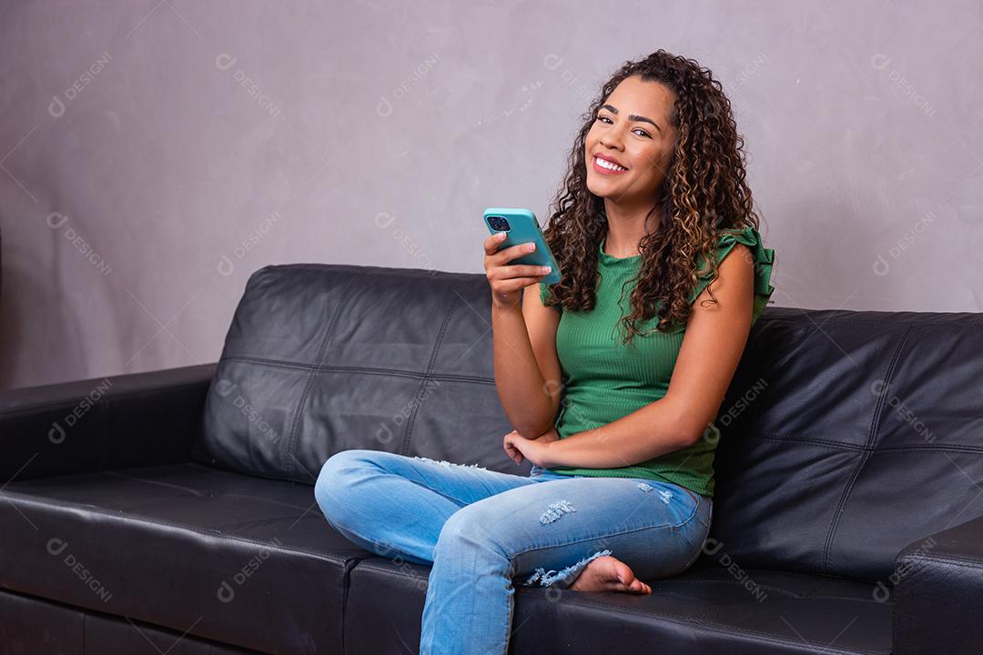 Uma jovem bastante africana descansa no sofá, usa um sorriso de telefone inteligente, olha para o bate-papo