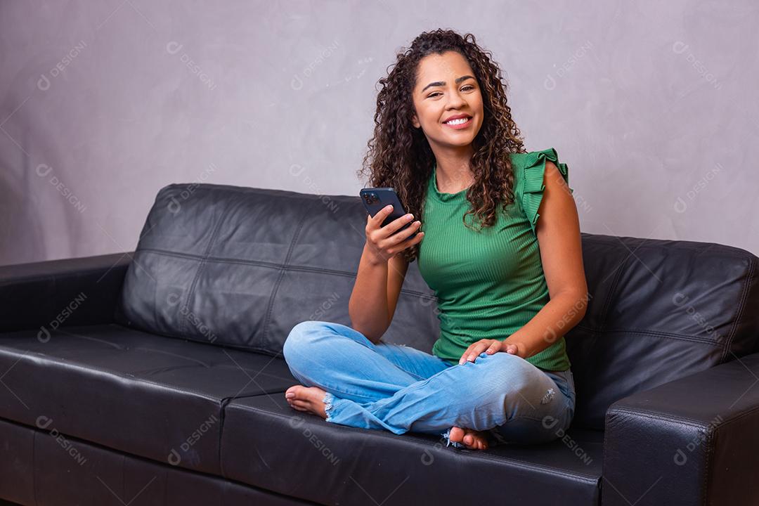 Uma jovem bastante africana descansa no sofá, usa um sorriso de telefone inteligente, olha para o bate-papo