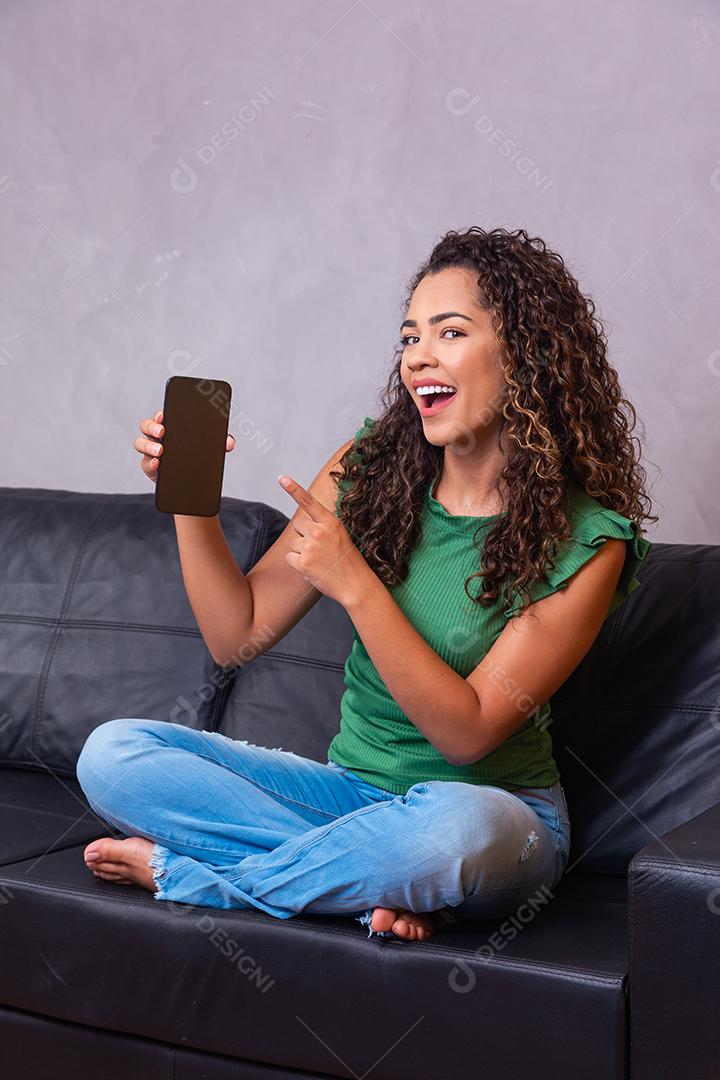 Mulher afro sentada no sofá segurando o celular com tela branca