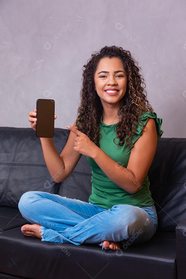 Mulher afro sentada no sofá segurando o celular com tela branca com espaço livre para texto.