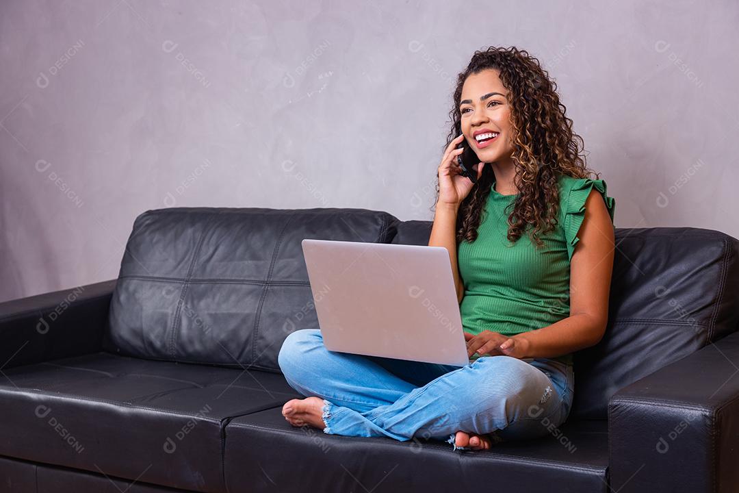 Mulher afro-americana milenar sorridente falando ao telefone usando laptop no escritório em casa