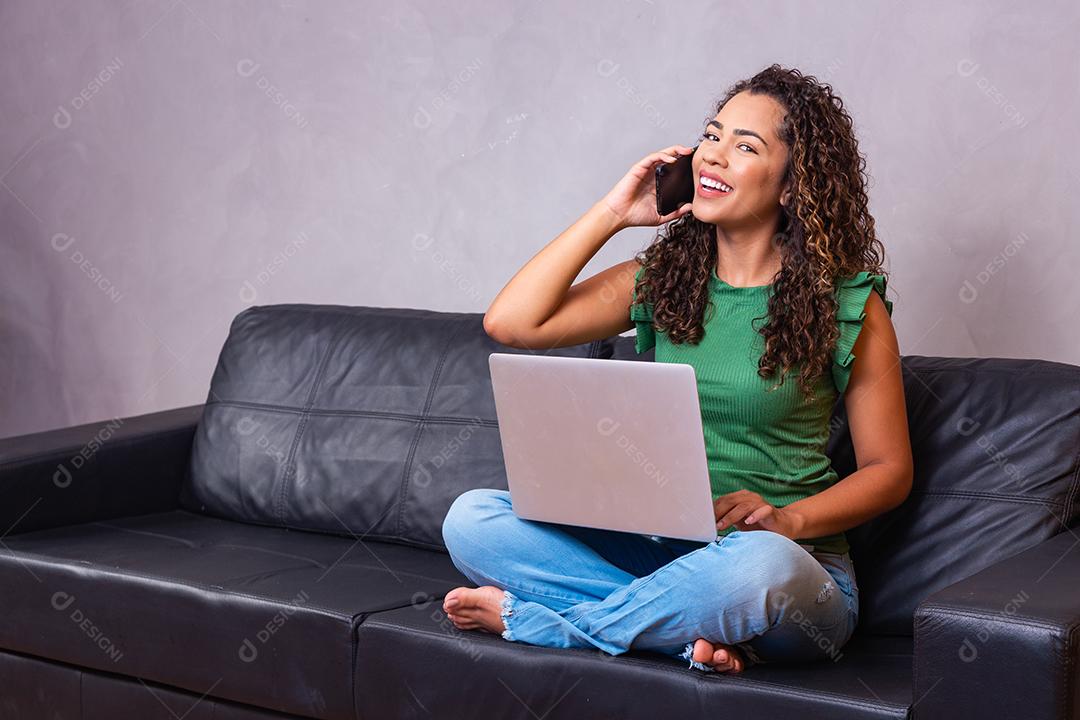 Mulher afro-americana milenar sorridente falando ao telefone usando laptop no escritório em casa