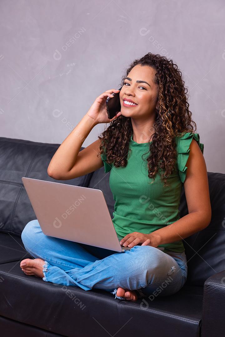 Mulher afro-americana milenar sorridente falando ao telefone usando laptop no escritório em casa