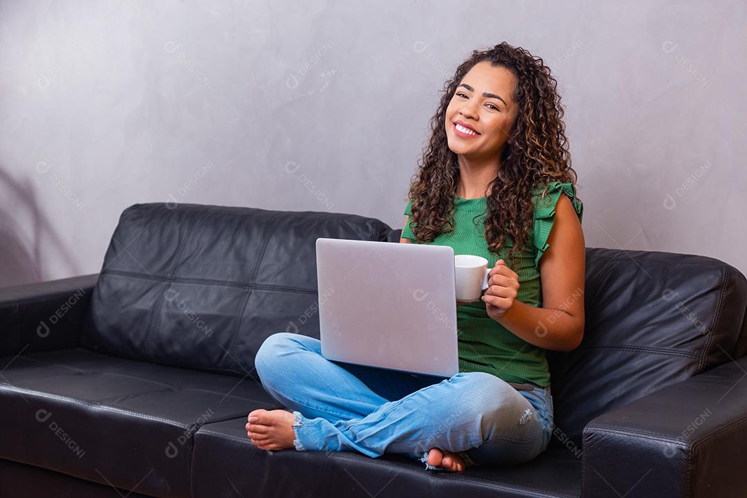 Sorrindo jovem afro usando laptop, sentado no sofá em casa, linda garota fazendo compras ou conversando