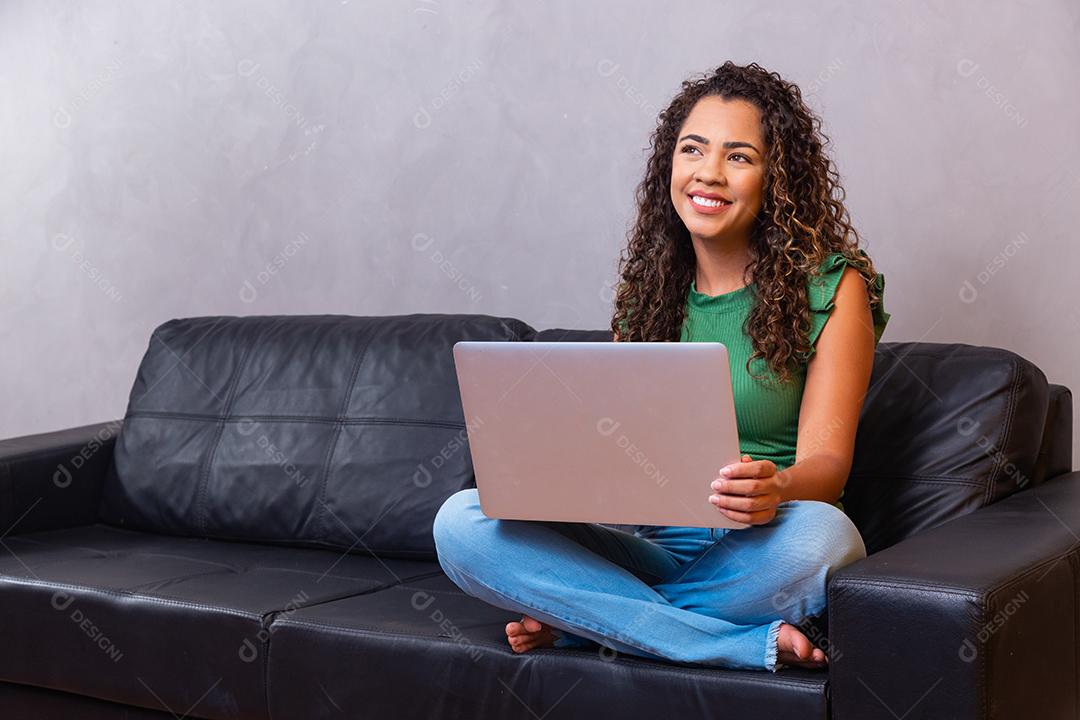 Sorrindo jovem afro usando laptop, sentado no sofá em casa, linda garota fazendo compras ou conversando