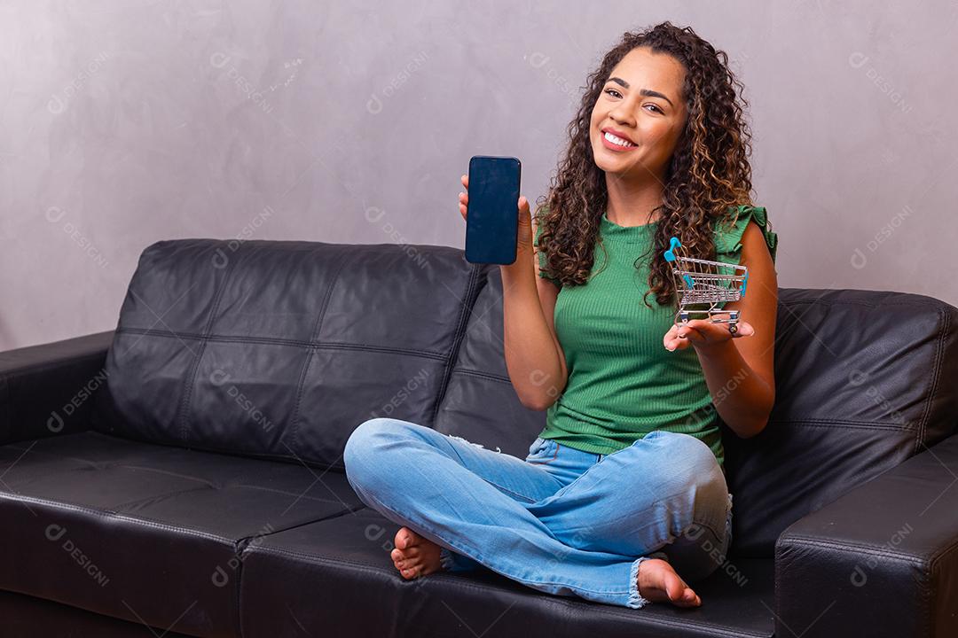 Mulher segurando celular sentado no sofá segurando carrinho de supermecado