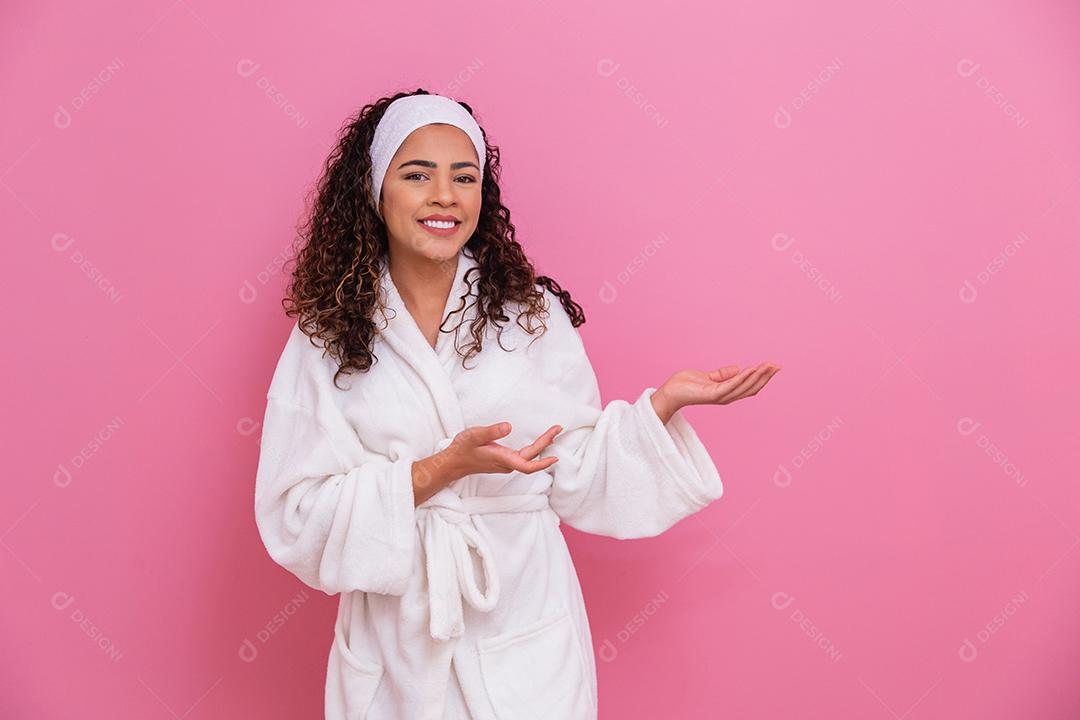 Linda mulher afro, vestida com um roupão de banho, apontando para o lado no fundo rosa