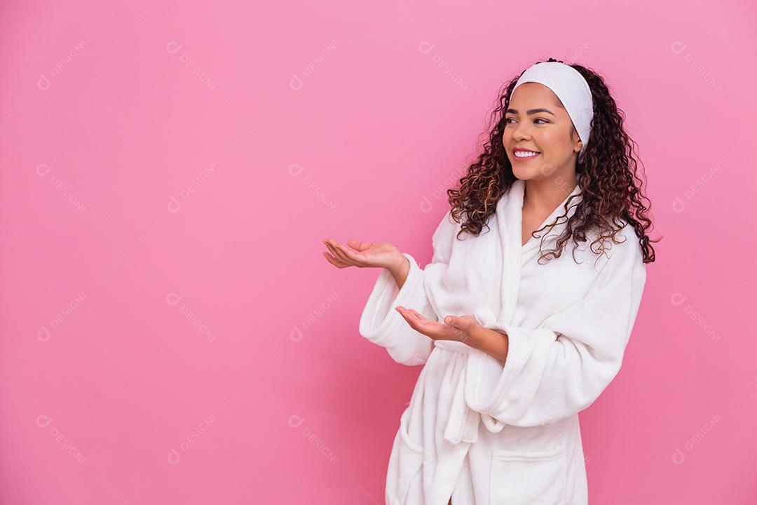 Linda mulher afro, vestida com um roupão de banho, apontando para o lado no fundo rosa
