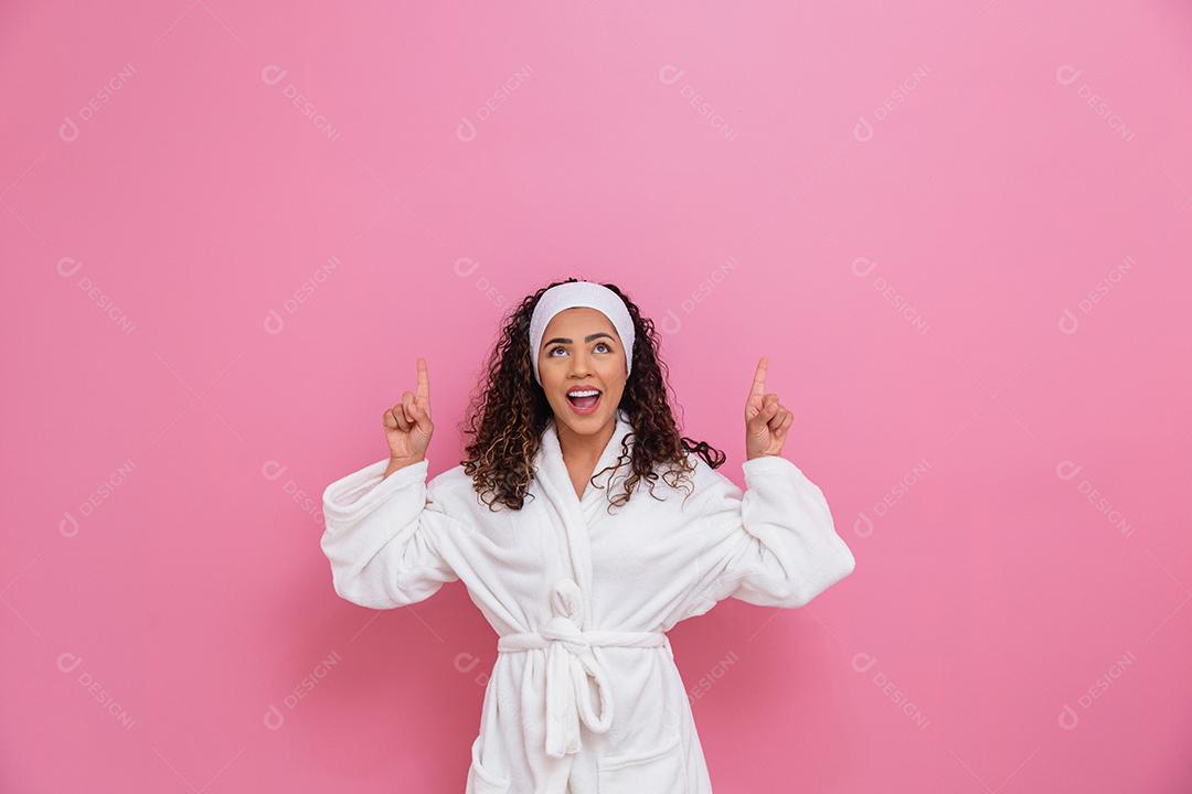 Linda mulher afro vestida com um roupão branco apontando para cima sorrindo.