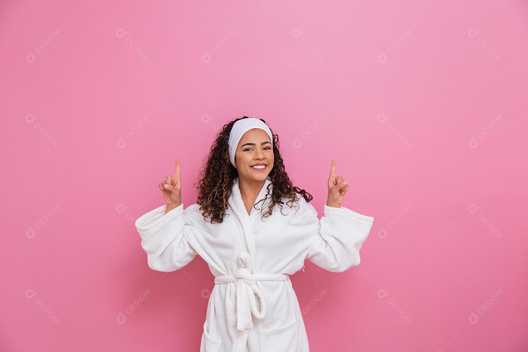 Linda mulher afro vestida com um roupão branco apontando para cima sorrindo.