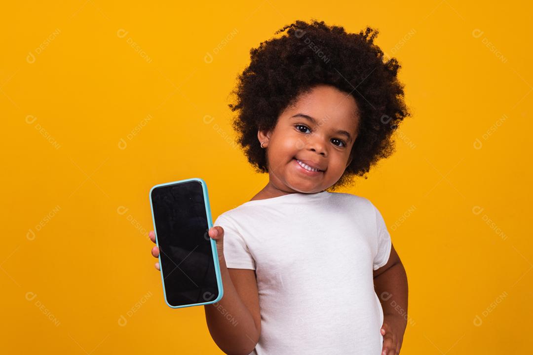 menina pequena criança afro segurando um telefone e apontando isolado em fundo amarelo.