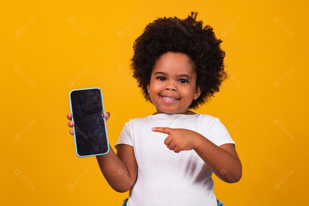menina pequena criança afro segurando um telefone e apontando isolado em fundo amarelo.