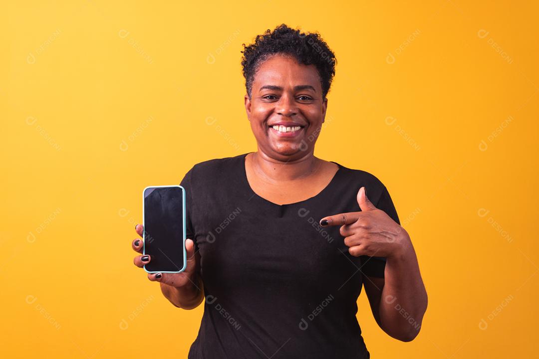 Closeup de sorridente mulher afro segurando smartphone com tela preta em branco na mão em fundo amarelo.