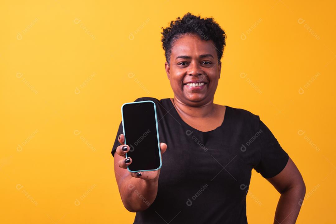 Closeup de sorridente mulher afro segurando smartphone com tela preta em branco na mão em fundo amarelo.